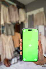 A close-up of a hand holding a smartphone with a green screen for mockup use, with blurred clothing...