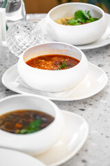 A close-up of three stylish bowls of soup placed on a modern table. One bowl features a vibrant red soup, while the others contain darker broths, garnished with herbs.