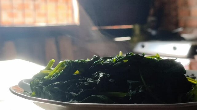 A plate of steaming boiled cassava leaves in a rural kitchen with a window in the background with sunlight coming in.