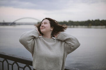 Portrait of happy young woman near river on city bridge in cold cloudy weather.