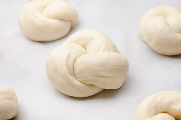 Raw yeast bread buns on parchment paper. Braided knot rolls or small yeast bread knots resting or second rise. Swiss butter bread called Zopf or Challah. Sunday bread. Selective focus.