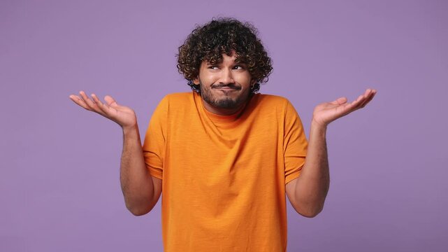 Hesitant young Indian man he wear orange t-shirt casual clothes feels doubtful spreading hands say oops ouch oh omg i am so sorry isolated on plain pastel light purple background. Lifestyle concept