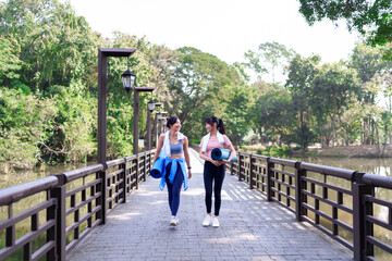Two young Asian women walking on a wooden bridge in a park, cooling down after an outdoor workout