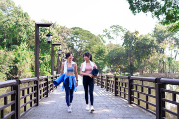 Two young Asian women walking on a wooden bridge in a park, cooling down after an outdoor workout