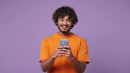 Happy smiling calm young Indian man wear orange t-shirt casual clothes hold use mobile cell phone typing browsing chatting send sms isolated on plain pastel light purple background. Lifestyle concept
