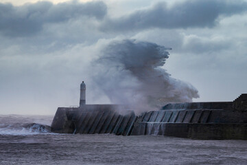A huge winter storm hitting the coast of the UK. Giant waves crash into a breakwater, with a lighthouse. The sky is grey and moody