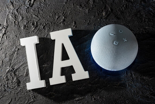 Letras tridimensionales blancas con el texto IA junto a un dispositivo Amazon Echo Dot, reflejando la integraci&oacute;n de la inteligencia artificial en el hogar, sobre fondo oscuro.