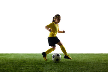 Obraz premium Girl, child in yellow jersey and black shorts playing soccer on grassy surface against white studio background. Training session. Concept of sport, childhood, motion, competition, fun, activity.
