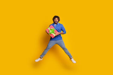 Fototapeta na wymiar Happy young man jumps holding a gift box with a green ribbon against vibrant yellow background, radiating joy and excitement