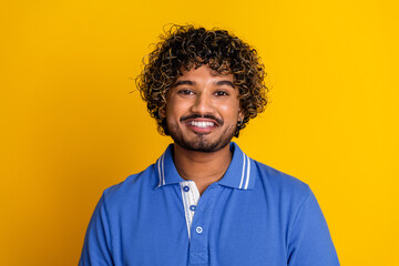 Smiling young man with curly hair wearing a blue polo shirt standing against yellow background