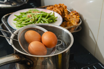 A metal strainer holds three freshly boiled eggs above a steaming pot of water, home cook preparation