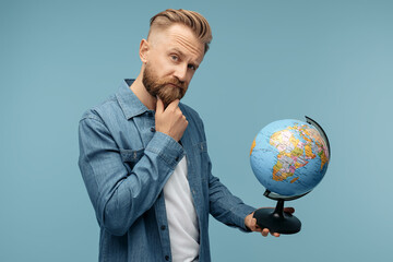 Thinking bearded hipster blonde young man taking holding world globe over blue background