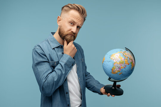 Thinking bearded hipster blonde young man taking holding world globe over blue background