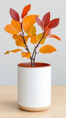 Autumnal branch with vibrant red and orange leaves in a white cylindrical pot on a light wooden surface. Simple, minimalist home decor.