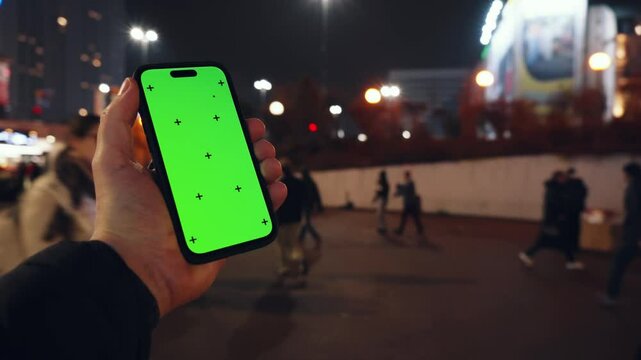 Pov view male hand holding modern smartphone keylight green screen gadget in street at night, camera locked on cell phone chroma key display, bokeh city illumination in evening, copy space