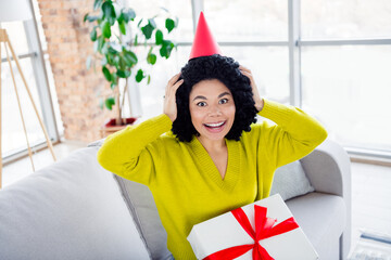 Happy young woman with a gift box and party hat celebrating indoors in a bright and stylish living...
