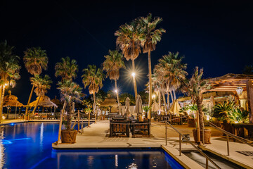 A luxurious tropical resort at night with a pool, palm trees, warm lights, and cozy seating,...