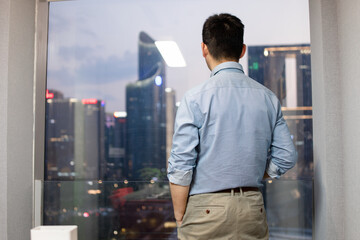 The man is standing by the window of the office at night.