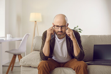 Serious confused puzzled man sitting on sofa with laptop computer at home, thinking, trying to...