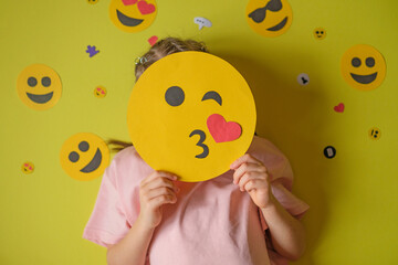 The child holds smiley face that sends an air kiss. The girl is surrounded by a lot of emoticons. The concept of problem of influence of social networks and social media on psychology of adolescents