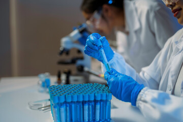 Scientist examining blue liquid test tube while colleague analyzing sample through microscope during medical research in sterile laboratory setting