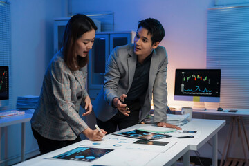 Business colleagues reviewing financial charts and graphs, analyzing market data spread across desk in sleek, dimly lit corporate workspace late in workday