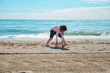 A child enjoys a sunny day playing on a sandy beach near the ocean waves under a sky with scattered clouds.
