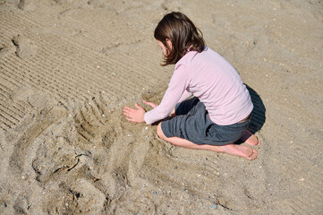 Child immersed in drawing in the sandy shore, capturing the essence of playful summer moments in...