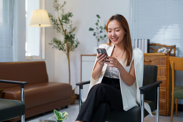 Professional Asian businesswoman working on smartphone, seated in sleek office lobby with leather sofa, floor lamp, and indoor greenery, radiating confidence