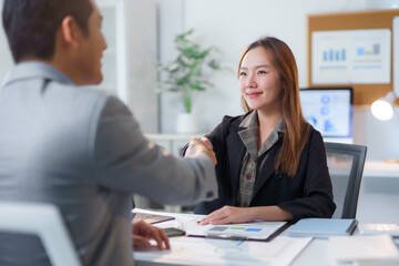 Obraz premium Two asian business people shaking hands over a desk with documents, laptop and financial charts, celebrating successful deal or agreement in modern office