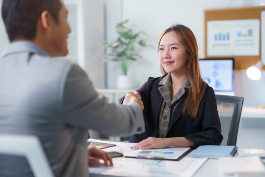Two asian business people shaking hands over a desk with documents, laptop and financial charts, celebrating successful deal or agreement in modern office