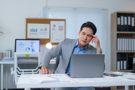Exhausted Asian professional suffering migraine, gripping head while working on laptop at office desk, experiencing intense work related stress and physical strain from prolonged employment