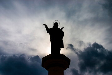 Backlit silhouette of a Jesus Christ statue with open arms, set against a dramatic cloudy sky, evokes feelings of faith and spirituality