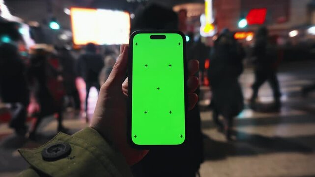 Pov looking at male hand holding smartphone keylight green screen gadget in street at night, man walking crowded crosswalk with phone chroma key display, bokeh modern city illumination in evening