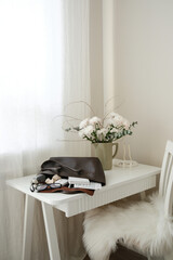 A stylish white desk adorned with vibrant fresh flowers, chocalate handbag with accessories of woman: notebooks, keyboard, bottle of water, glasses on white desk in light room.