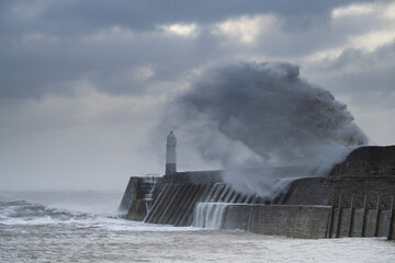 A huge winter storm hitting the coast of the UK. Giant waves crash into a breakwater, with a lighthouse. The sky is grey and moody © parkerspics