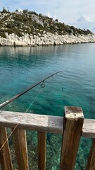 Fototapeta premium A fishing rod resting on a wooden pier overlooking a calm turquoise sea, with a rocky coastline and green hills in the background. A peaceful fishing scene capturing the beauty of nature and outdoor r