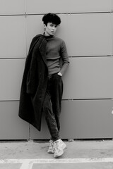 Fashionable black and white portrait of a man posing against a gray building with a coat draped...