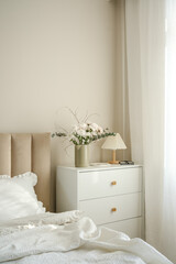 A serene bedroom with a stylish bed, plush pillows, and fresh flowers on white dresser artfully arranged in the room.