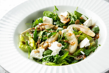 A fresh salad featuring mixed greens, sliced pears, crumbled feta cheese, pumpkin seeds, and a light dressing, presented in a white dish.