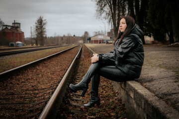 Fototapeta premium A woman in a black leather outfit and high-heeled boots sits near railway tracks, gazing into the distance. The overcast sky and rustic setting create a moody, contemplative atmosphere.
