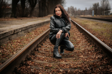 A woman in a black leather outfit and boots crouches on railway tracks, gazing thoughtfully. The...