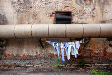 The picture shows a courtyard in the city with a large brick wall and a pipe. Beneath this pipe, there is a rope with various dresses hanging up to dry. Depressing picture