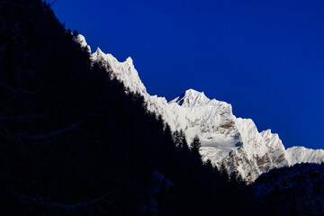 snow-covered slope with dangerous peaks is illuminated by the morning sun against the blue sky. In front of the snow-covered peaks, you can see a dark forest located in the shadow of nearest mountains