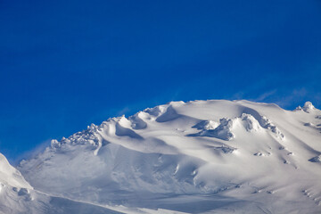 The snow-covered surface of the mountainside. The top of this mountain is exposed to strong winds that blow snowflakes away like dust against the blue sky
