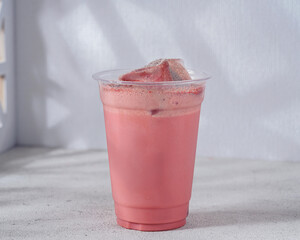 A refreshing iced strawberry milk drink served in a clear plastic cup with ice cubes. The creamy pink beverage is infused with strawberry flavor and topped with a dusting of cocoa powder for a delight