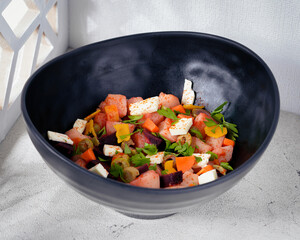 A vibrant mixed vegetable and cheese salad served in a stylish black bowl. The dish includes diced potatoes, carrots, beets, olives, and feta cheese, garnished with fresh parsley and a sprinkle of pap