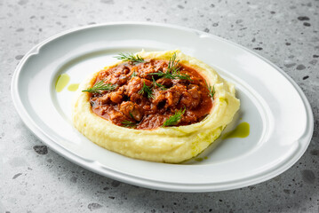 A sleek plate features creamy mashed potatoes topped with a savory stew, garnished with fresh dill. The dish offers a warm, inviting presentation against a textured gray background.