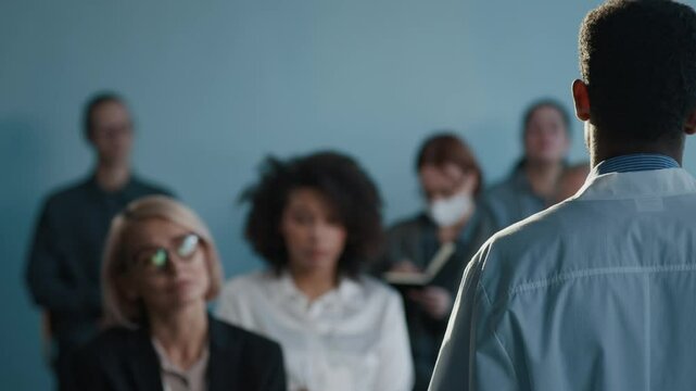 Rear view of unrecognizable Black male scientist wearing lab coat giving lecture to group of business people