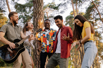 Multiracial group of people, bearded hipster man playing guitar and friends dancing, singing and...